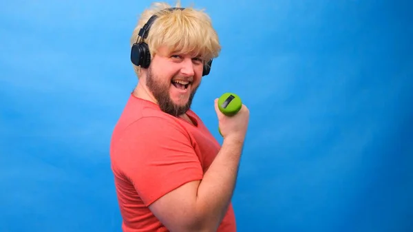 Hombre gordo divertido freaky en una peluca y una camiseta rosa con pesas haciendo ejercicios sobre un fondo azul . —  Fotos de Stock