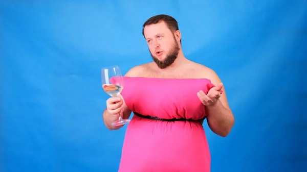 Divertido barbudo con un vestido rosa hecho de almohadas bebe vino de una copa sobre un fondo azul. Cuarentena loca. Moda 2020. Ponte una almohada. Desafío 2020 debido al aislamiento de la casa — Foto de Stock