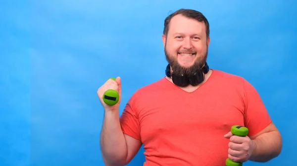 Engraçado homem gordo esquisito em uma peruca e uma camiseta rosa com halteres fazendo exercícios em um fundo azul . — Fotografia de Stock