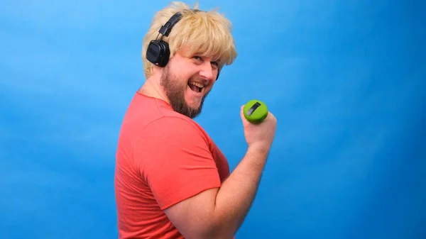 Vicces kövér ember furcsa parókában és rózsaszín pólóban súlyzókkal gyakorolja a kék hátteret.. — Stock Fotó