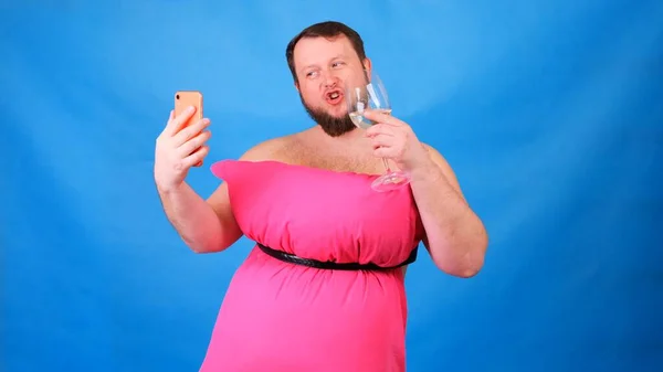 Cara barbudo engraçado em um vestido rosa feito de travesseiros com um copo de vinho faz selfie em um fundo azul. Quarentena maluca. Limpeza de casa engraçada. Moda 2020. Põe uma almofada. Desafio 2020 devido a — Fotografia de Stock