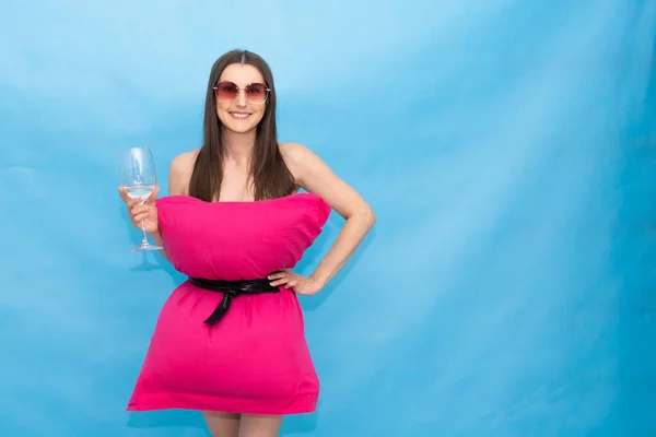 Ευτυχισμένη γυναίκα με ροζ μαξιλάρι φόρεμα και γυαλιά ηλίου κρατώντας ένα ποτήρι σαμπάνια σε ένα μπλε φόντο για την τρελή πρόκληση Pillow οφείλεται στην ύπαρξη στο σπίτι στην απομόνωση. Μοντέρνο κορίτσι. Μαξιλάρι κλήση λόγω — Φωτογραφία Αρχείου