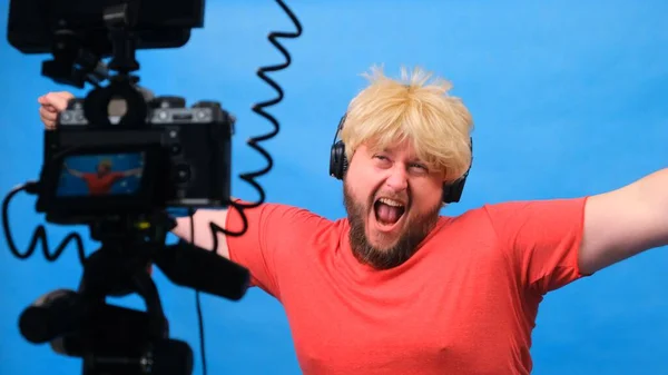 Homem gordo estranho em uma peruca e uma camiseta rosa faz um blog de vídeo contra um fundo azul . — Fotografia de Stock