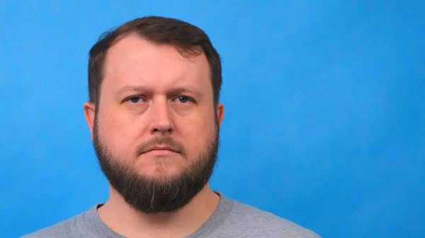 Portrait of a sad bearded man in a gray t-shirt singing and talking to the camera on a blue background. — Stock Photo, Image