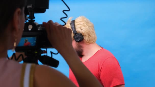 Freaky dikke man in een pruik en een roze t-shirt maakt een video blog tegen een blauwe achtergrond. — Stockvideo