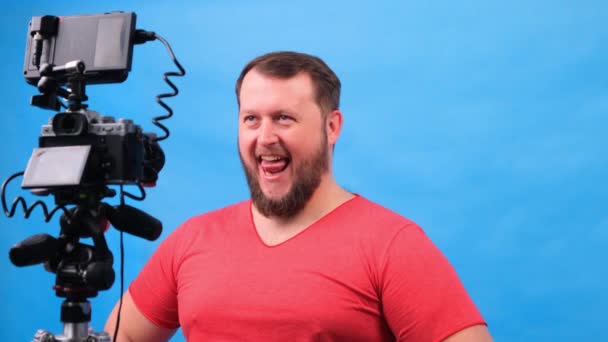 Jonge dikke man in een roze t-shirt maakt een video blog tegen een blauwe achtergrond. — Stockvideo