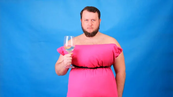 Rolig skäggig kille i rosa klänning gjord av kuddar är ledsen med ett glas vin på en blå bakgrund. Galen karantän. Rolig städning. Mode 2020. Sätt på dig en kudde. Utmaning 2020 på grund av huset — Stockfoto