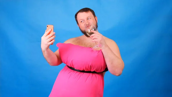 Lustiger bärtiger Typ in einem rosafarbenen Kleid aus Kissen mit einem Glas Wein macht Selfie auf blauem Hintergrund. Verrückte Quarantäne. Lustige Hausreinigung. Mode 2020. Auf ein Kissen legen. Herausforderung 2020 — Stockfoto