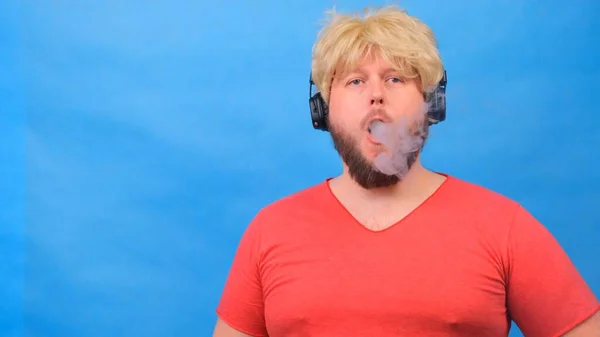 Странный толстяк в парике и розовая футболка в наушниках выдыхает сигаретный дым, курит парик на синем фоне. — стоковое фото