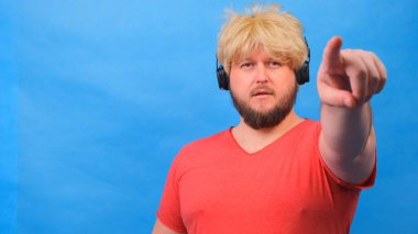 Peruklu ve kulaklıklı pembe tişörtlü ucube şişman adam mavi arka planda sana doğru bir parmak gösteriyor..