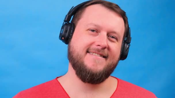 Bearded funny cute dikke man in een roze T-shirt luistert naar muziek en danst op een blauwe achtergrond, close-up, kopieer ruimte. — Stockvideo