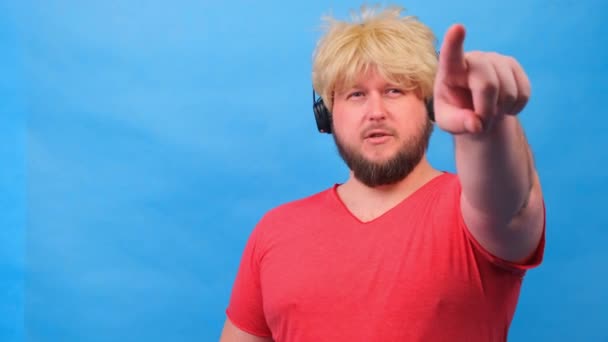 Hombre gordo raro en una peluca y una camiseta rosa en los auriculares muestra un dedo en la cámara en usted contra un fondo azul . — Vídeos de Stock