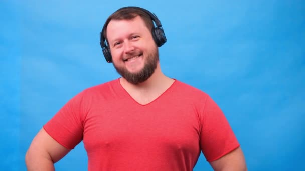 Barbudo divertido sonriente lindo gordo hombre en una camiseta rosa escucha música y baila sobre un fondo azul, primer plano, espacio de copia . — Vídeo de stock