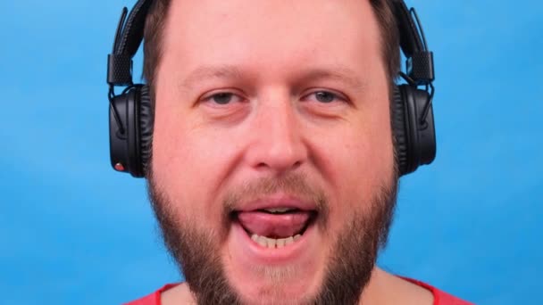 Vousatý legrační roztomilý tlustý muž v růžovém tričku poslouchá hudbu a tance a ukazuje svůj jazyk na modrém pozadí, zblízka, kopírovací prostor. — Stock video
