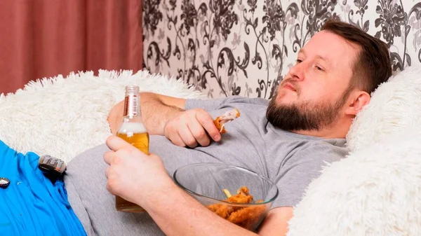 Kövér ember sört iszik és egészségtelen csirkeszárnyakat eszik, unatkozik a tévé előtt a kanapén. Az alultápláltság, az otthoni karantén, az alkoholizmus fogalma. — Stock Fotó