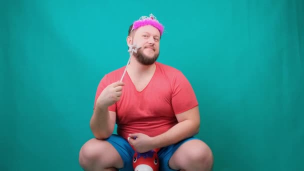 Schattige bebaarde freaky man in een roze T-shirt met een diadeem op zijn hoofd droomt van het berijden van een eenhoorn met een toverstaf in zijn hand. Een grappige tovenaar grap te maken en een wens te vervullen — Stockvideo
