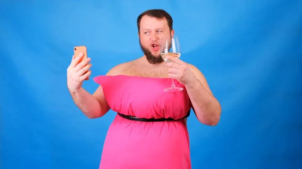 Rolig skäggig kille i rosa klänning gjord av kuddar med ett glas vin gör selfie på en blå bakgrund. Galen karantän. Rolig städning. Mode 2020. Sätt på dig en kudde. Utmaning 2020 på grund av — Stockfoto