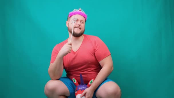 Schattige bebaarde freaky man in een roze T-shirt met een diadeem op zijn hoofd droomt van het berijden van een eenhoorn met een toverstaf in zijn hand. Een grappige tovenaar grap te maken en een wens te vervullen — Stockvideo