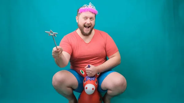 Cute bearded freaky man in a pink T-shirt with a diadem on his head dreams of riding a unicorn with a magic wand in his hand. A funny wizard joke to make and fulfill a wish — Stock Photo, Image