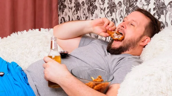 Hombre gordo bebe cerveza y come alimentos poco saludables alas de pollo, aburrido delante de la perspectiva de la televisión en el sofá. El concepto de desnutrición, cuarentena en casa, alcoholismo . — Foto de Stock