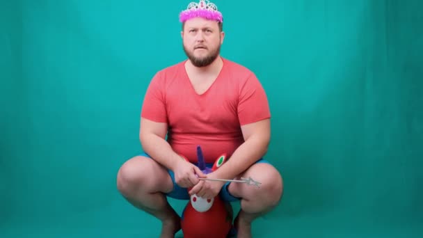 Handsome bearded freaky man in a pink T-shirt with a deadema on his head is saddened riding a unicorn with a magic wand in his hand. A funny wizard joke to make and fulfill a wish. — Stock Video