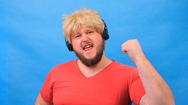 Peruklu, pembe tişörtlü garip şişman adam kulaklıkla müzik dinliyor ve mavi arka planda dans ediyor.