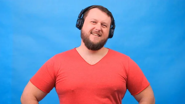 Γενειάδα αστείο χαμογελαστό χαριτωμένο λίπος άνθρωπος σε ένα ροζ T-shirt ακούει μουσική και χορούς σε ένα μπλε φόντο, κοντινό πλάνο, αντίγραφο χώρο. — Φωτογραφία Αρχείου