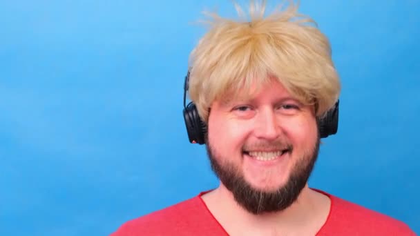 Hombre gordo raro en una peluca y una camiseta rosa en sus manos escucha música en los auriculares y baila sobre un fondo azul — Vídeos de Stock