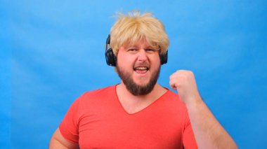 Peruklu, pembe tişörtlü garip şişman adam kulaklıkla müzik dinliyor ve mavi arka planda dans ediyor.