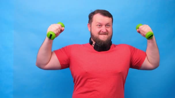 Fat fancy man in a pink t-shirt with dumbbells funny doing exercises on a blue background. — Stock Video