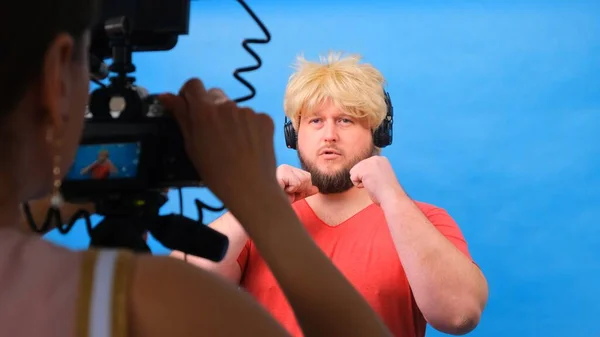 Homem gordo estranho em uma peruca e uma camiseta rosa faz um blog de vídeo contra um fundo azul . — Fotografia de Stock