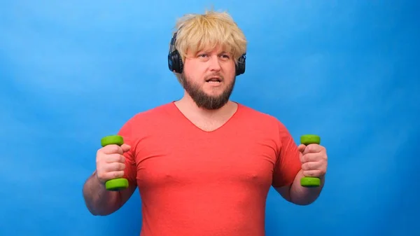 Funny fat man freaky in a wig and a pink t-shirt with dumbbells doing exercises on a blue background. — Stock Photo, Image