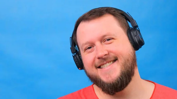 Bearded funny smiling cute fat man in a pink T-shirt listens to music and dances on a blue background, close-up, copy space. — Stock Photo, Image