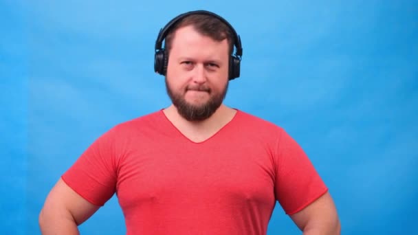 Hombre gordo carismático alegre en auriculares aislados y camiseta rosa sobre un fondo azul. escuchar música, cantar y bailar — Vídeos de Stock