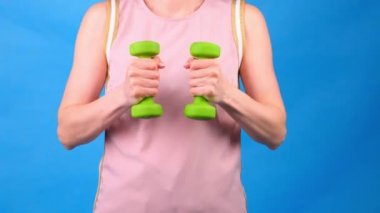 Elinde dambıllar olan pembe spor takım elbiseli bir kadın mavi arka planda egzersiz yapıyor. Spor, diyet ve kilo kaybı kavramı.