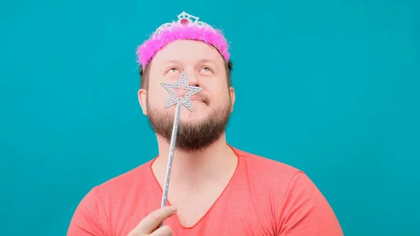 Divertido barbudo hombre raro en una camiseta rosa con un deadema en la cabeza está soñando con una varita mágica en la mano. Un divertido chiste de mago para hacer y cumplir un deseo . — Foto de Stock