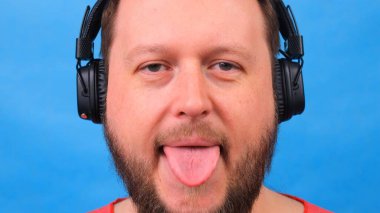 Sakallı, pembe tişörtlü komik şişman adam müzik dinler, dans eder ve dilini mavi arka planda gösterir..