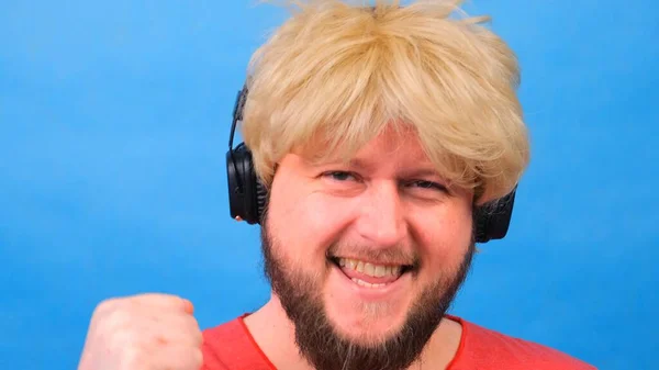 freaky fat man in a wig and a pink T-shirt in his hands listens to music on headphones and dances on a blue background
