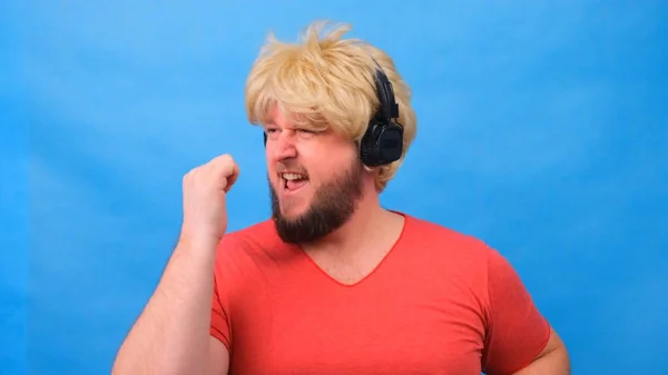 Freaky fat man in a wig and a pink T-shirt in his hands listens to music on headphones and dances on a blue background — Stock Photo, Image