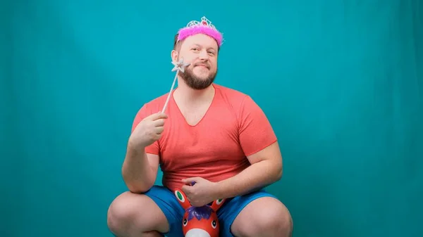 一个戴着粉色T恤，头戴头戴皇冠的可爱的胡子怪胎男人，梦想着骑着一只手里拿着魔杖的独角兽。一个有趣的巫师笑话来实现一个愿望 — 图库照片