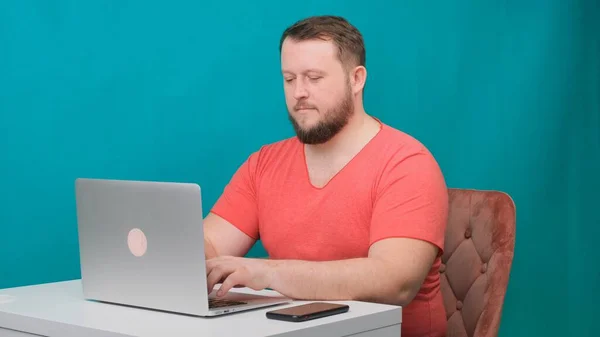 Il giovane uomo d'affari felice in una t-shirt rosa lavora su un computer portatile su uno schermo verde. Ritratto di un uomo parlante che guarda nel suo portatile. Uomo che lavora alla sua scrivania in ufficio. — Foto Stock