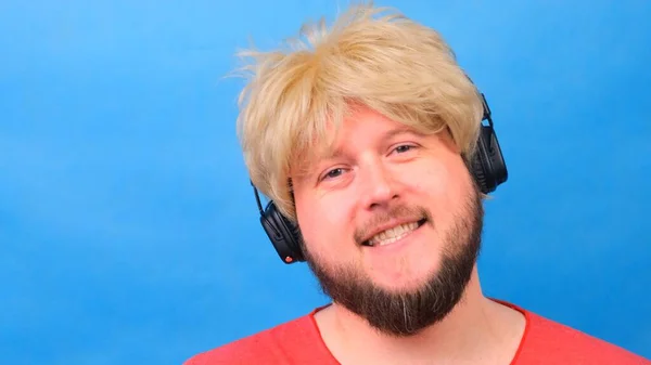 Freaky fat man in a wig and a pink T-shirt in his hands listens to music on headphones and dances on a blue background — Stock Photo, Image