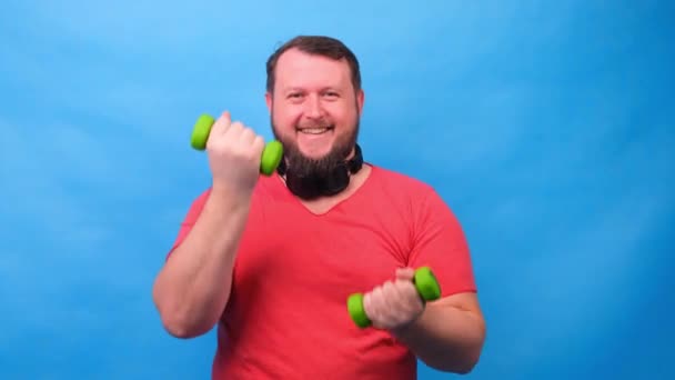 Gros fantaisie homme dans un t-shirt rose avec haltères drôle faire des exercices sur un fond bleu. — Video