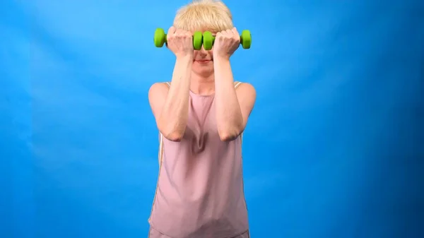Смешная женщина в парике с белыми волосами с зелеными гантелями в руках делает упражнения на синем фоне — стоковое фото