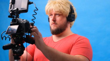 Peruklu ve pembe tişörtlü garip şişman adam mavi arka planda video bloğu yapıyor..