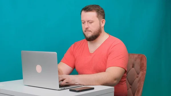 Молодий щасливий бізнесмен у рожевій футболці працює на ноутбуці на зеленому екрані. Портрет чоловіка, який розмовляє, дивлячись у свій ноутбук. Чоловік працює за своїм столом в офісі . — стокове фото