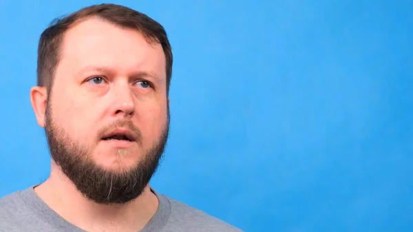 Retrato de un hombre barbudo triste con una camiseta gris cantando y hablando con la cámara sobre un fondo azul . —  Fotos de Stock