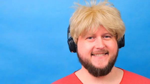 Lachender, dicker Mann mit Perücke und rosa T-Shirt in der Hand hört Musik über Kopfhörer und tanzt auf blauem Hintergrund — Stockfoto