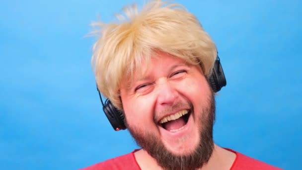 Ridendo uomo grasso strambo con una parrucca e una maglietta rosa tra le mani ascolta musica su cuffie e balli su uno sfondo blu — Video Stock