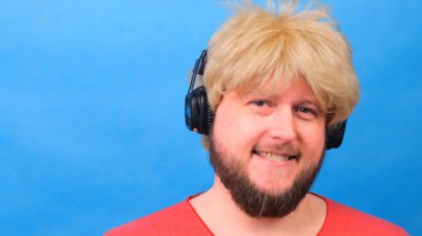 Peruklu, pembe tişörtlü garip şişman adam kulaklıkla müzik dinliyor ve mavi arka planda dans ediyor.